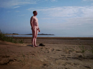 A nude man stands alone on a sandy beach, gazing at the tranquil sea under a vast, blue sky dotted with soft clouds. The golden sand stretches around him, with beach grasses swaying gently in the light breeze, creating a serene, idyllic atmosphere.