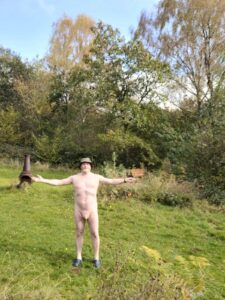 A man standing nude in a serene outdoor setting, surrounded by lush greenery and vibrant trees. He wears a sun hat and stands with arms outstretched, embracing the natural beauty around him. The scene is peaceful, capturing a moment of freedom in a tranquil landscape.