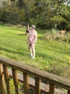 A man stands naked in a grassy garden, wearing a sun hat and blue flip-flops. He is turned slightly to his left, surrounded by trees and shrubs under a clear sky. The wooden railing of a porch is visible in the foreground.