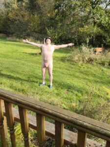 A man stands confidently in a lush green garden, arms outstretched as if embracing the sunlight. He is nude, wearing only a wide-brimmed hat and blue shoes, with a backdrop of trees and overgrown grass, creating a sense of freedom and spontaneity in nature.