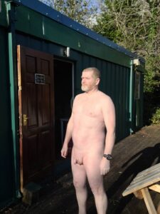 A man stands outside a green metal cabin, illuminated by sunlight. He has short hair and a beard, wearing a watch, while his skin is exposed to the elements. The background features lush greenery, adding a natural feel to the scene.