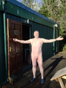 A man standing outside a green cabin with his arms outstretched, appearing relaxed and confident. He is nude, with a light complexion and short hair, wearing dark blue slippers. The entrance of the cabin is open, revealing a sunny day with lush trees in the background.