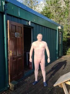 A man stands outside a green shipping container-like structure, appearing fully nude except for blue slippers. The scene is brightly lit by natural sunlight, surrounded by trees and greenery. An open door behind him reveals a glimpse of the interior.