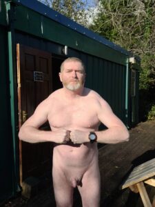 A man standing confidently outside a wooden building, with hands on hips, showcasing a strong physique. He has short grey hair, a beard, and is wearing only a wristwatch. The scene is set in a natural, leafy environment under clear blue skies.