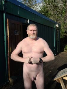 A bare-chested man stands confidently outside a wooden building, with a shadow of trees in the background. He has a light beard and is wearing a watch on his right wrist. The sun casts a warm glow on his skin, suggesting a bright day.