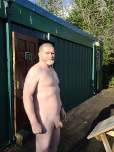 A man stands outdoors, slightly to the side, with a serious expression. He is nude, revealing light skin and a rugged physique, amid a green and serene setting. Behind him is a green container building with a closed door, surrounded by trees under bright sunlight.