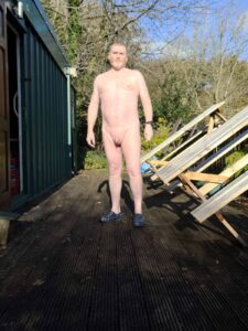 A man standing in a natural setting, naked, on a textured wooden deck. Sunlight casts shadows, highlighting his relaxed posture. Surrounding him are wooden structures tilted against the deck and foliage in the background, creating a serene outdoor atmosphere.
