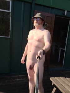 A man taking a selfie outdoors, standing confidently in the sunlight. He is wearing sunglasses and a wide-brimmed hat, showcasing his bare skin against a backdrop of a green cabin and wooden decking. The scene exudes a sense of freedom and leisure.
