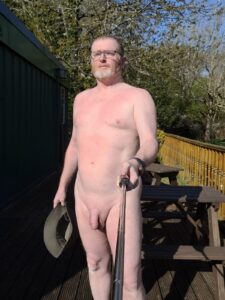 A middle-aged man with grey hair and glasses poses for a selfie outdoors, standing on a wooden deck in bright sunlight. He is nude, holding a hat in one hand while the other extends the selfie stick. Background features lush greenery and a wooden railing.