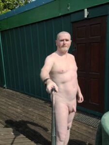 A man standing outdoors, holding a selfie stick, captured in a nude pose. He stands on a wooden deck in front of a green metal structure, with a closed wooden door visible behind him. Natural sunlight illuminates the scene, casting soft shadows.