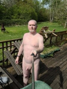 A middle-aged man stands naked on a wooden deck, holding a selfie stick, with a calm expression on his face. Behind him, a lush green garden stretches out, dotted with trees and a few wooden chairs. The sunlight bathes the scene in a warm glow, enhancing the tranquil atmosphere.