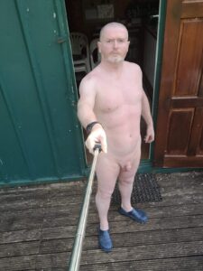 A man stands outside a wooden door, holding a selfie stick. He is nude, with a confident posture. He has a shaved head and a short beard, wearing only blue clogs. The rustic green wall behind him complements the natural setting, creating a striking contrast.
