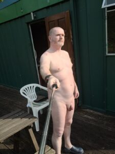 A man stands outdoors, confidently posing for a selfie while holding a selfie stick. He is nude, with light skin and short hair, facing slightly to the side. In the background, a wooden deck features a white plastic chair and a green metal building, with sunlight illuminating the scene.