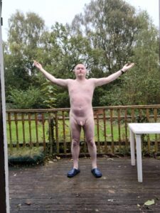A man standing outdoors on a wooden deck, with arms raised in a gesture of celebration or freedom. He is naked and wears dark slippers, and the background features lush greenery and soft, overcast skies, creating a tranquil natural setting.