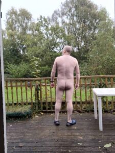 A man standing nude on a wooden deck, facing away from the camera towards a lush garden. He has a bald head and appears relaxed, wearing only blue slippers. Trees and greenery surround the area, creating a tranquil outdoor scene.