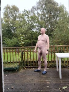 A man stands on a wooden deck, surrounded by lush greenery, with trees in the background. He is bald and has a neutral expression, wearing only blue slippers. The overcast sky adds a soft, diffused light to the scene, creating a calm atmosphere.