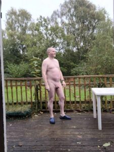 A man stands naked on a wooden deck surrounded by a lush garden, with tall trees and greenery in the background. He has a shaved head and is wearing blue slippers. The scene is set on a cloudy day, enhancing the natural, serene atmosphere.