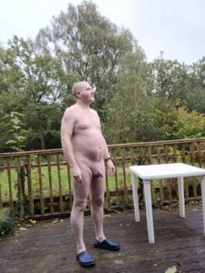 A man standing naked on a wooden deck, looking serene. He has a bald head and is wearing blue slippers. In the background, lush greenery with trees and shrubs creates a peaceful, natural setting. A white table sits nearby, adding to the outdoor atmosphere.