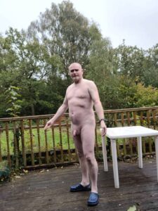 A man standing nude on a wooden terrace, surrounded by greenery. He has a light build and a shaved head, wearing blue slip-on shoes. The scene is set in a tranquil garden under overcast skies, with a white table nearby and the soft rustle of leaves in the background.