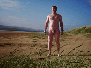 Naturist nudist nude naked penis man standing on beach with blue sky and landscape.