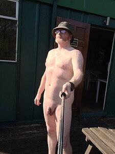 Nudist man with hat and sunglasses taking selfie outdoors.