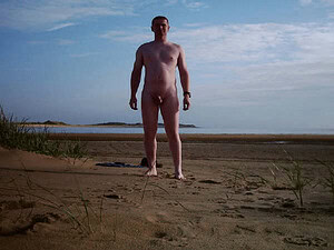 Naturist nudist nude naked penis man standing on beach with sky and sea background.