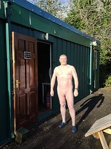 Nudist man standing outside a green cabin, fully nude, natural setting.
