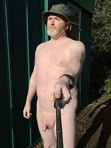 Nudist man outdoors with selfie stick, natural setting, relaxed pose, no clothing.