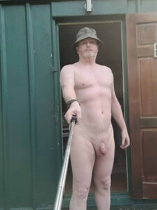 Nudist man with selfie stick, standing outdoors, no clothing, natural setting.