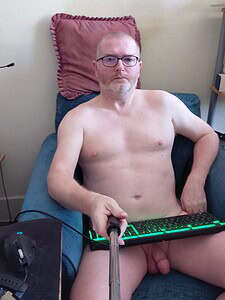 Naturist nudist nude naked penis man sitting on sofa with keyboard and selfie stick.